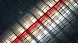 Overhead cinematic drone photograph of a vast industrial warehouse rooftop with a grid of frosted glass panels, illuminated from below with varying warm white and cool silver light. A sharp diagonal line of panels glows bright red, cutting across the frame. The upper-left quadrant is darker, and rainwater pools on the glass and in the seams, reflecting the light. The aesthetic is technical and futuristic.