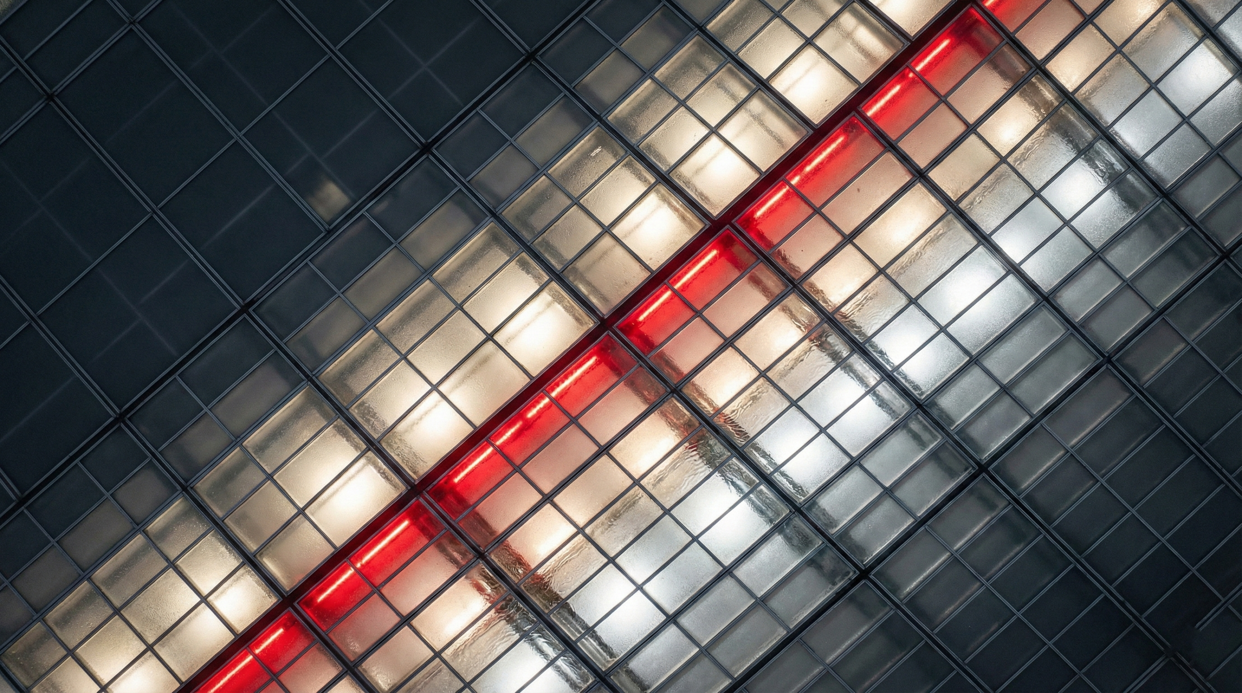 Overhead cinematic drone photograph of a vast industrial warehouse rooftop with a grid of frosted glass panels, illuminated from below with varying warm white and cool silver light. A sharp diagonal line of panels glows bright red, cutting across the frame. The upper-left quadrant is darker, and rainwater pools on the glass and in the seams, reflecting the light. The aesthetic is technical and futuristic.